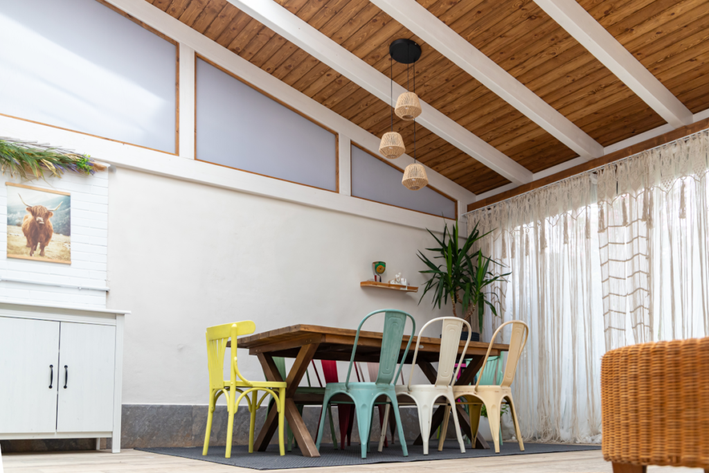 En la imagen se ve una mesa de comedor con sillas de colores en un porche de madera que amplia el salon