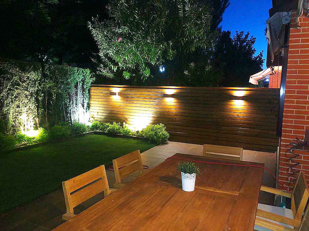 En la imagen se ve una valla de madera en un jardin con plantas iluminado por la noche